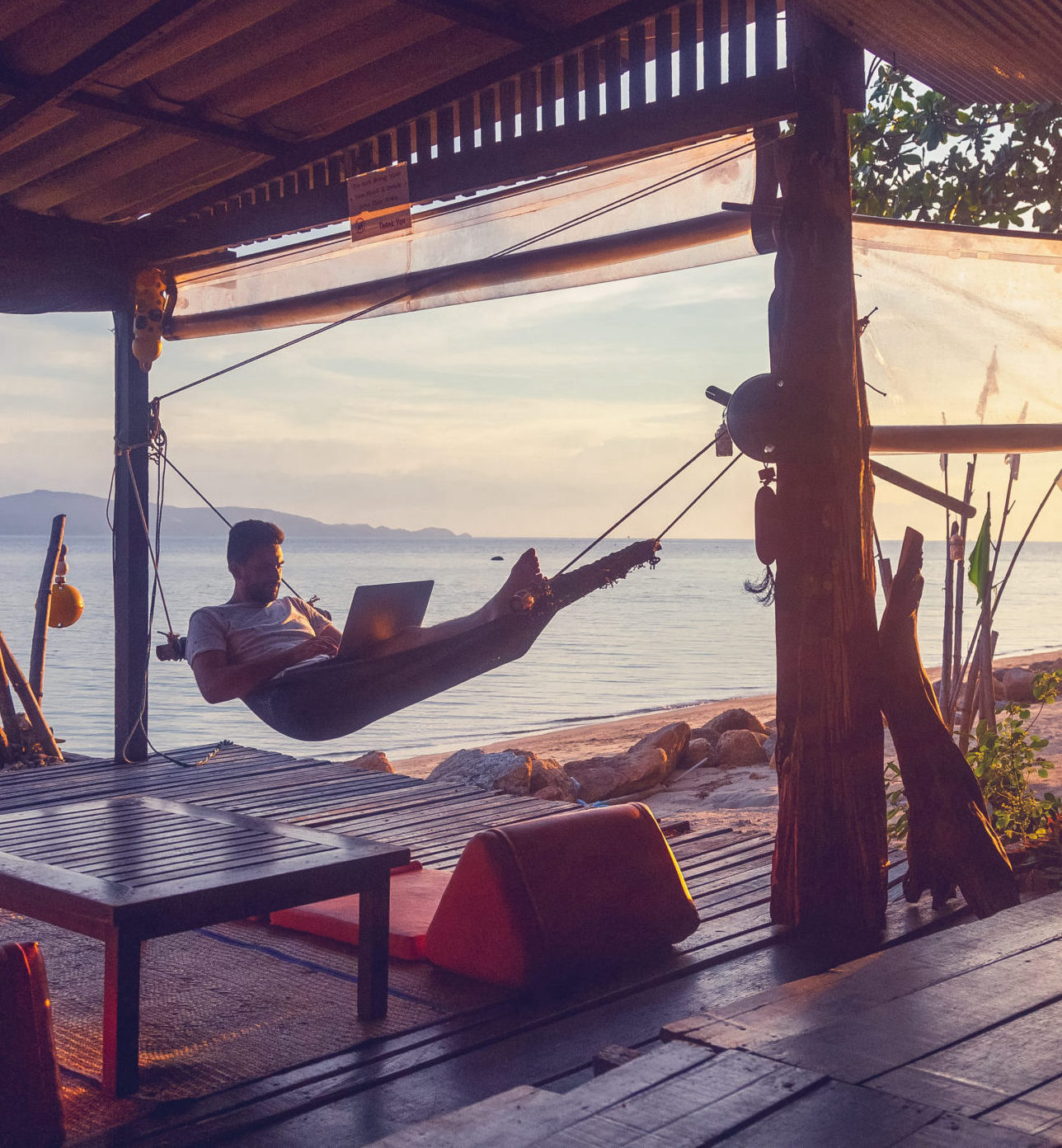 Young attractive guy a man with a beard in a hammock with a lapt Young attractive guy a man with a beard in a hammock with a laptop on the background of the sea and sunset, distant work, freelancer, blogger, vacation and travel
